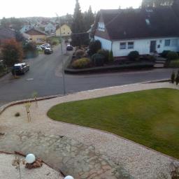 Ogrody Mościckiego - Widok z góry na zadbany ogród z trawnikiem, ścieżką z kamienia łupanego i żwiru, oraz niskie nasadzenia krzewów i drzewek iglastych, na tle domów jednorodzinnych.