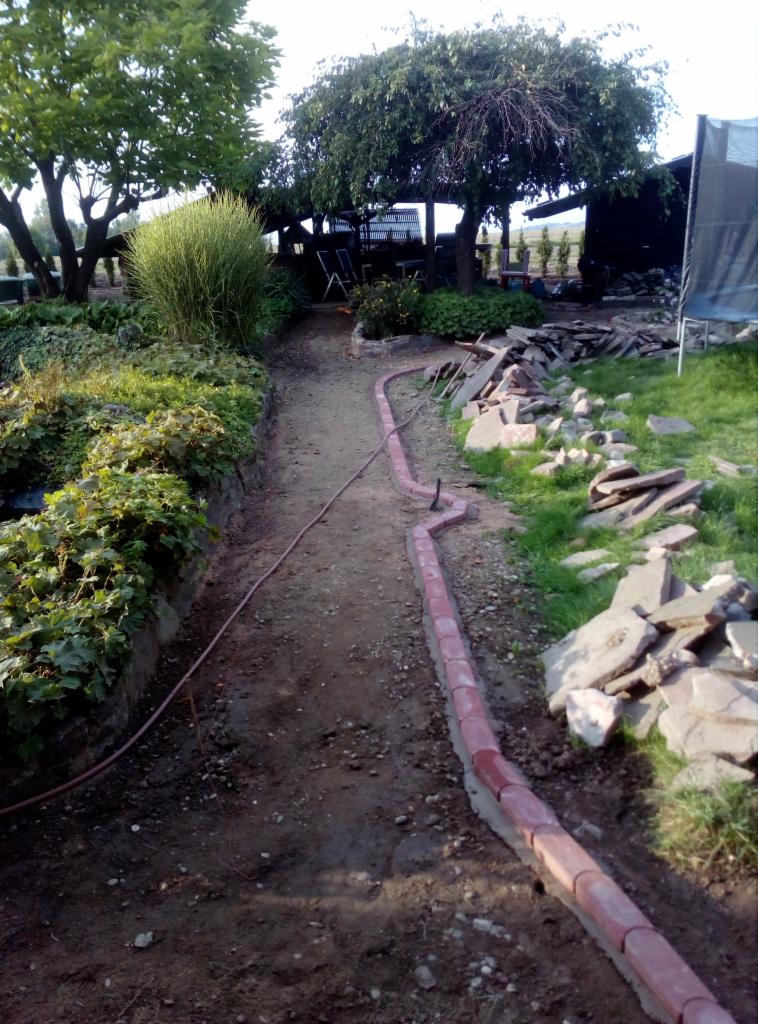 Budowa ścieżki w ogrodzie: ułożone obrzeża z czerwonej kostki, przygotowany grunt, kamienie do wyłożenia, widok na zieleń i altanę w tle.