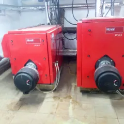 Dwa czerwone kotły gazowe Hoval Uno-3 z palnikami Giersch w kotłowni, widoczne rury i instalacje, płytki podłogowe z plamami.