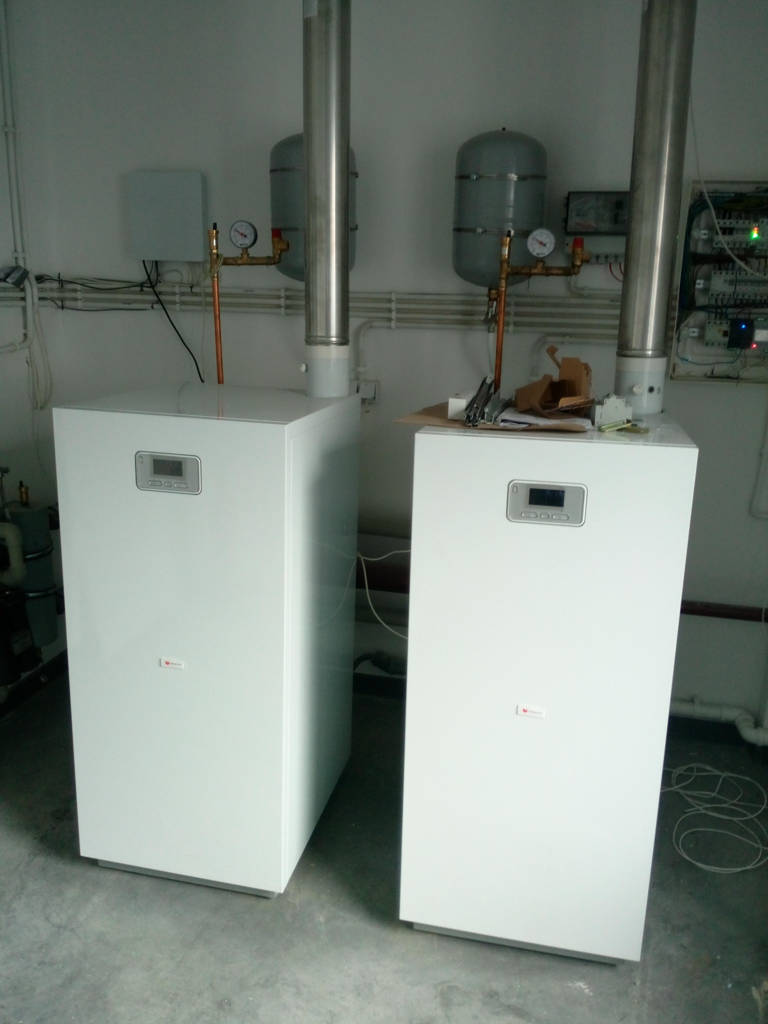 Dwa białe, nowoczesne kotły gazowe z pionowymi rurami odprowadzającymi spaliny, zainstalowane w pomieszczeniu technicznym z szarą podłogą i białymi ścianami, na których widoczne są instalacje.