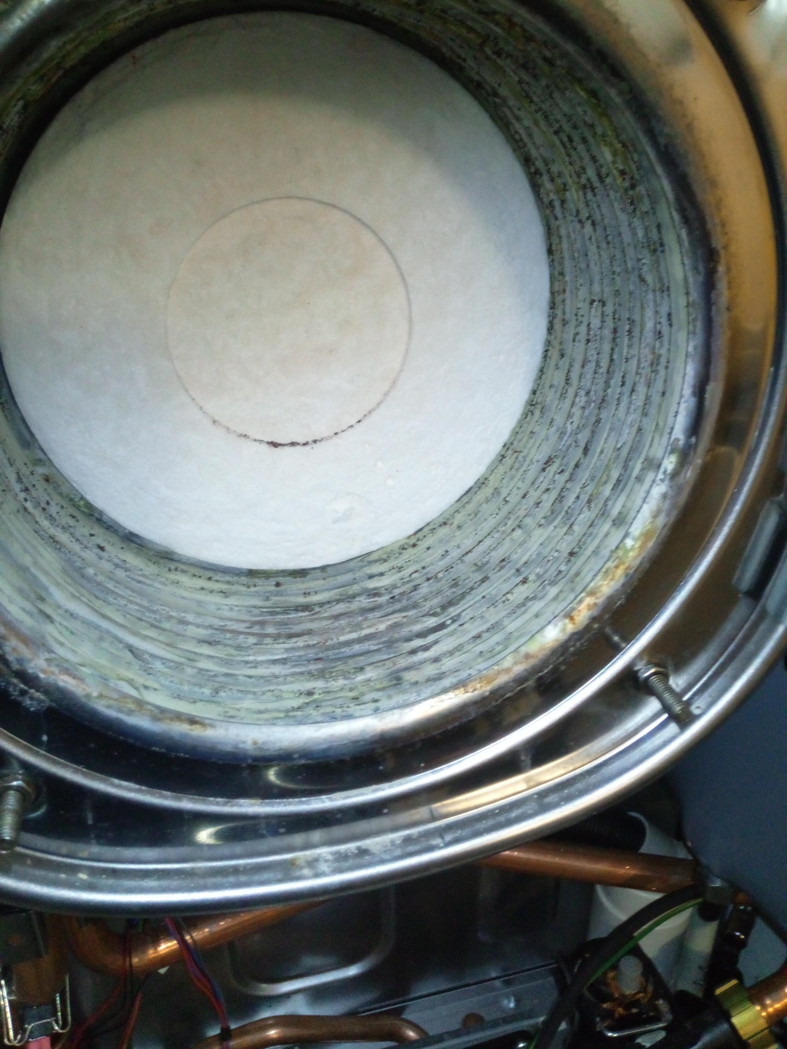 Wnętrze kotła gazowego z widocznym osadem na ściankach i elementem ceramicznym wewnątrz, z bliska widoczne miedziane rurki i śruby mocujące.