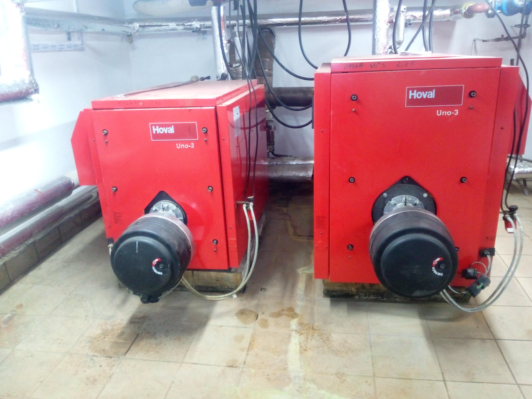 Dwa czerwone kotły gazowe Hoval Uno-3 z palnikami Giersch w kotłowni, widoczne rury i instalacje, płytki podłogowe z plamami.
