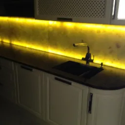 Nowoczesna kuchnia z podświetlanym panelem nad blatem, ciemnym blatem i białymi szafkami z frezowanymi frontami oraz chromowaną baterią.