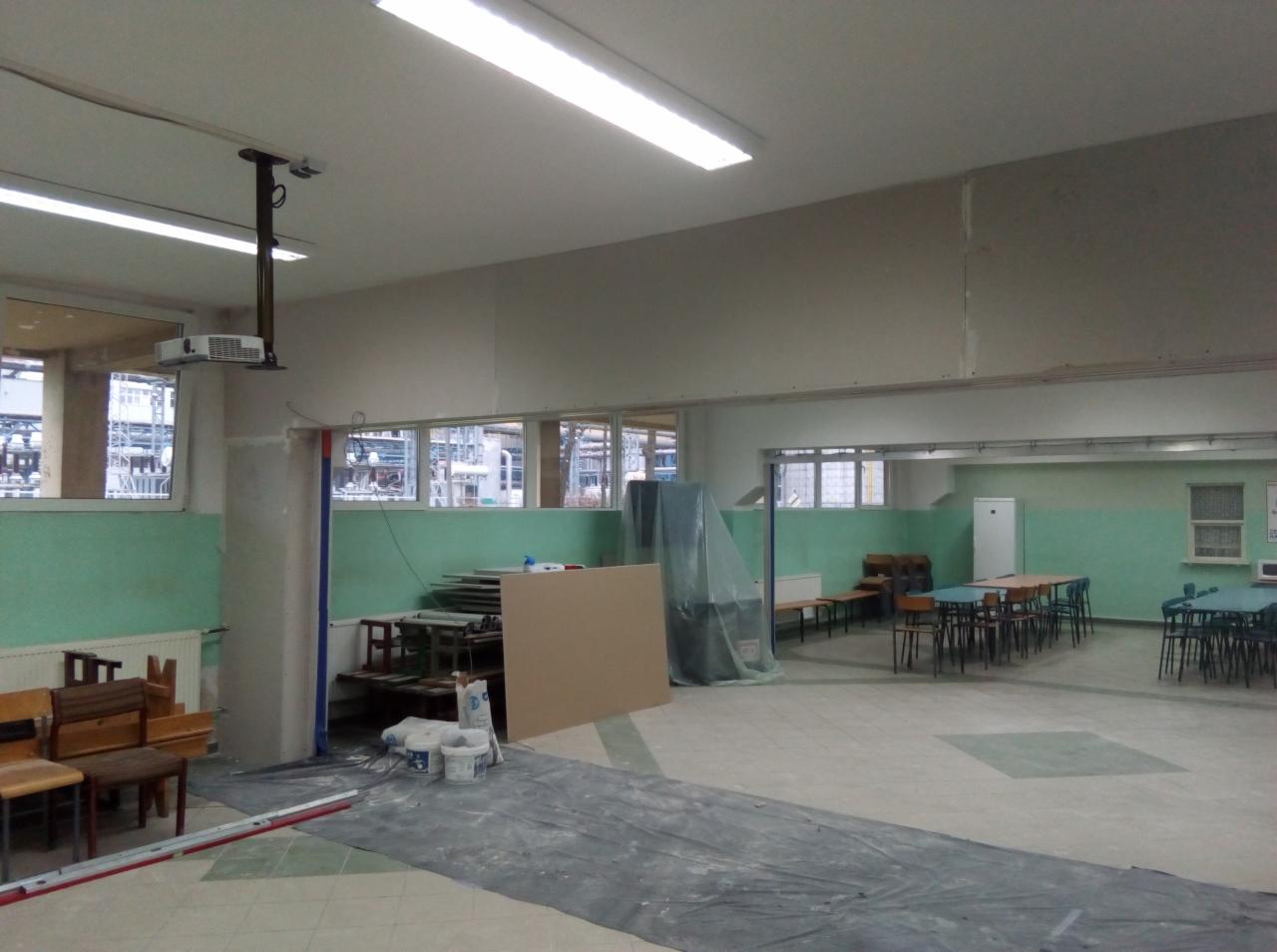 Wnętrze remontowanego pomieszczenia z widocznymi płytami gipsowo-kartonowymi na ścianie, przygotowanymi do dalszej obróbki, podłoga zabezpieczona folią malarską, a w tle ustawione stoły i krzesła.