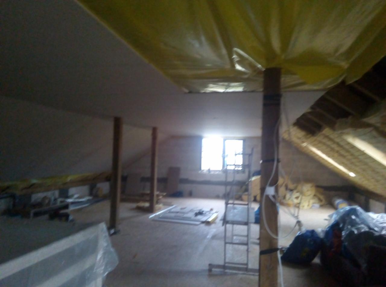 Poddasze w trakcie adaptacji, widoczne ocieplenie dachu, drewniane słupy podtrzymujące konstrukcję, okno i drabina w tle, folia zabezpieczająca na suficie.