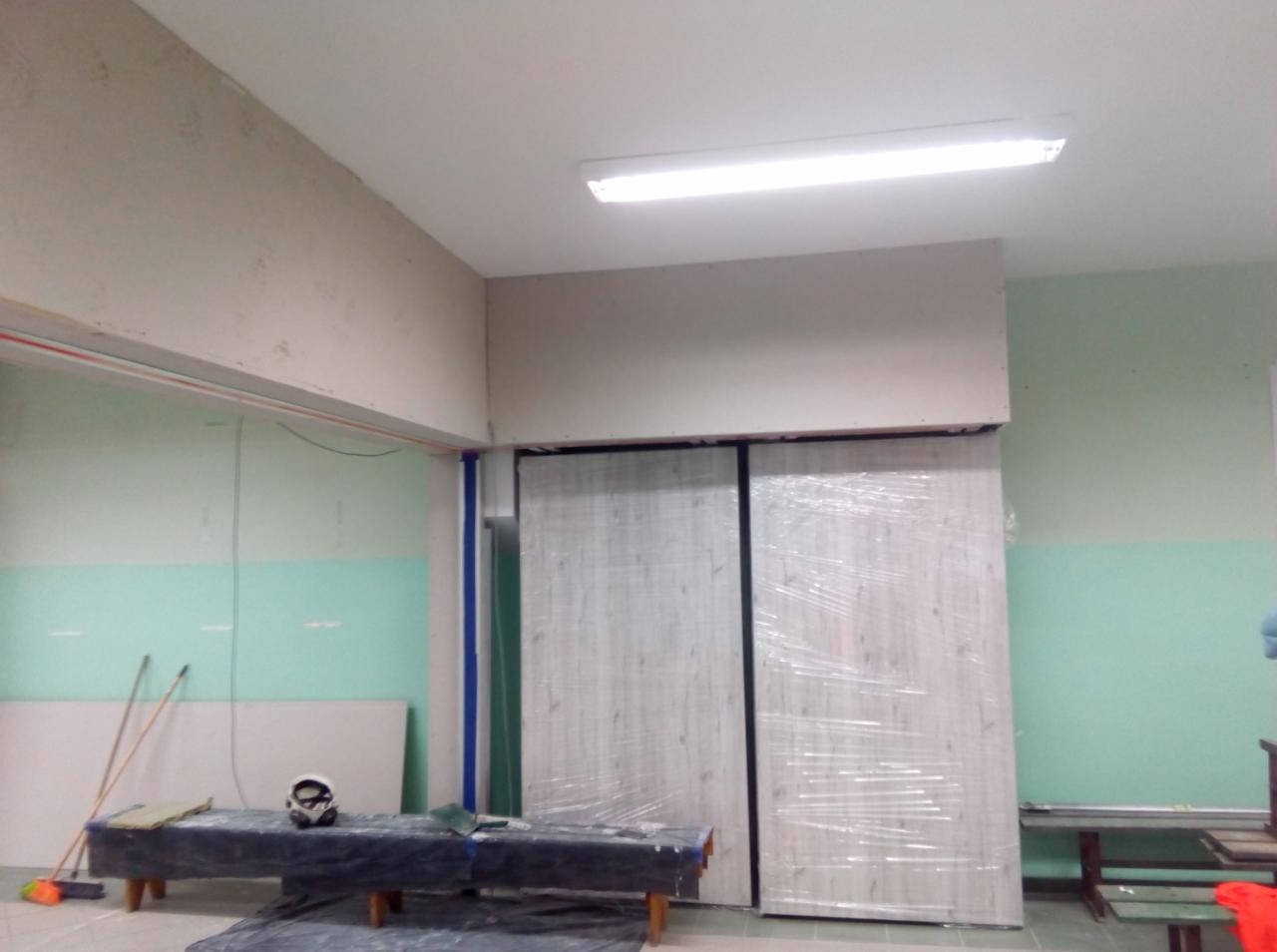 Przygotowanie do gładzi w pomieszczeniu: widoczne płyty gipsowo-kartonowe, zakryte folią drzwi przesuwne, maska ochronna i narzędzia na stole.