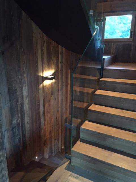 Drewniane schody z przeszkloną balustradą, oświetleniem ściennym i drewnianą okładziną ścienną, widok z góry na dół.