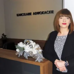 Kobieta w czarnym żakiecie stoi przy recepcji kancelarii adwokackiej, na której leży dekoracja kwiatowa ze srebrnymi elementami.