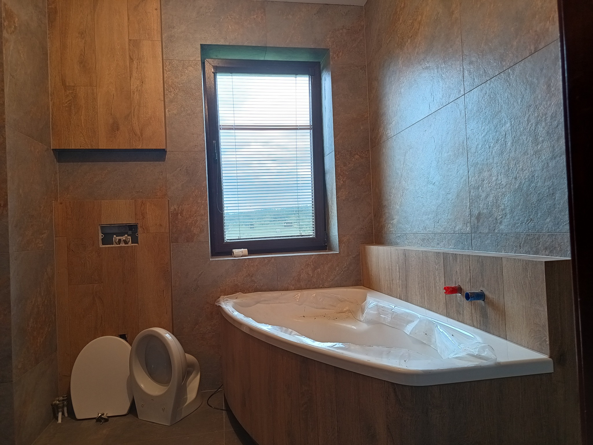 Łazienka w trakcie remontu: ułożone płytki imitujące drewno na ścianach, niezamontowana toaleta, wanna zabezpieczona folią, okno z żaluzjami.