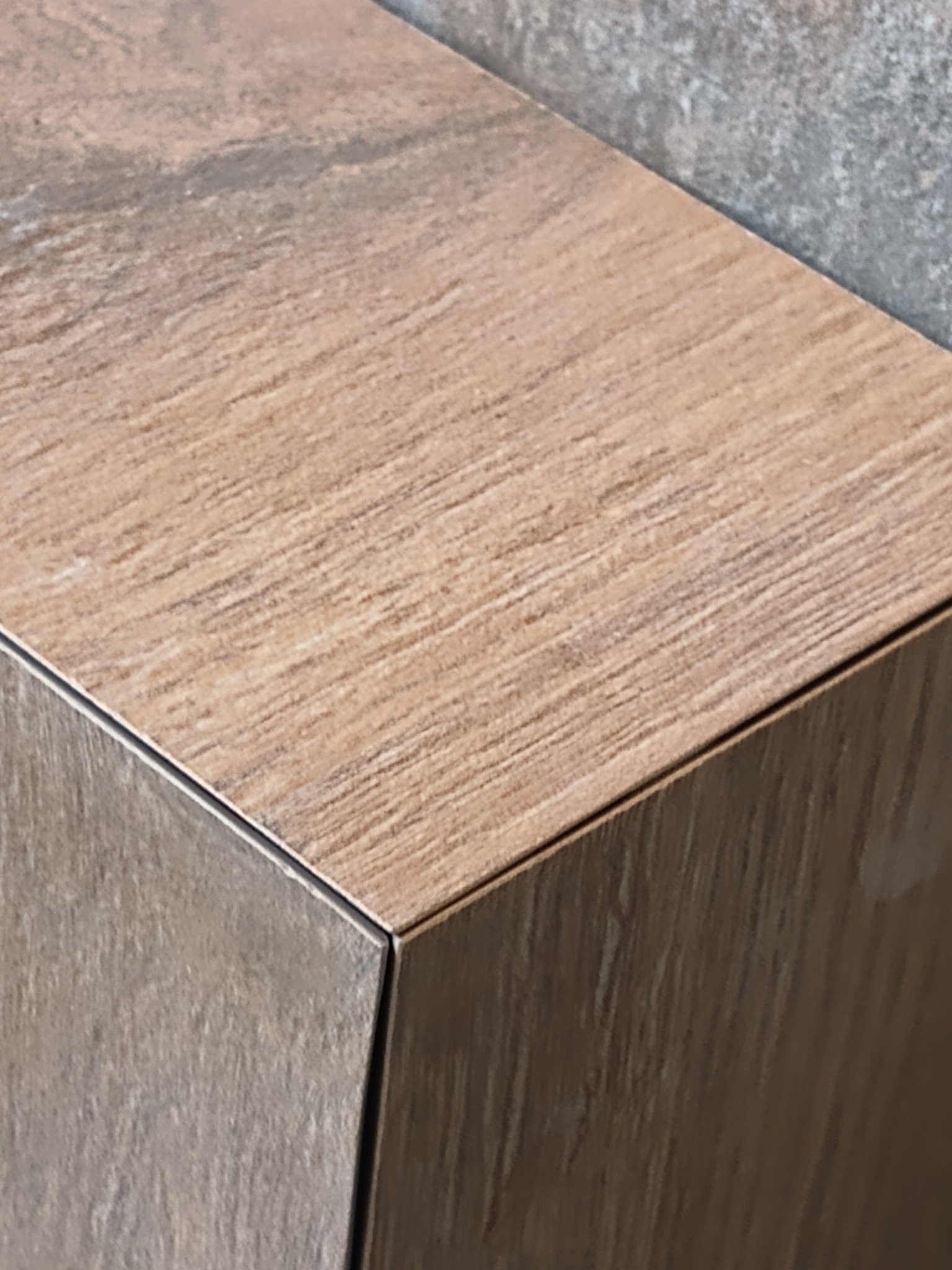 Detal narożnika obłożonego płytkami imitującymi drewno, z widoczną fugą i fragmentem szarej ściany w tle.