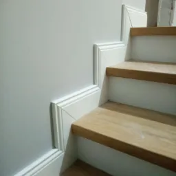 Szczegół wykończenia schodów: pomalowana na szaro ściana z białą listwą przypodłogową i drewniane stopnie z białymi podstopnicami.