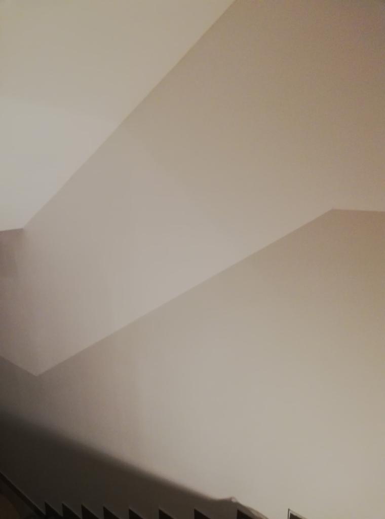 Świeżo wykończona, jasna ściana z widocznym załamaniem sufitu i fragmentem schodów w dolnej części kadru, ukazująca precyzyjne wykończenie powierzchni.