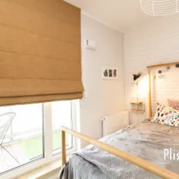 Beżowa, płócienna osłona okienna w stylu rzymskim, częściowo zwinięta, widoczna na tle jasnego pokoju z balkonem i białej ceglanej ściany.