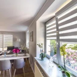 Salon z oknami przesłoniętymi roletami dzień noc, widok na ogród z kwitnącymi rododendronami, na parapecie storczyki w doniczkach.