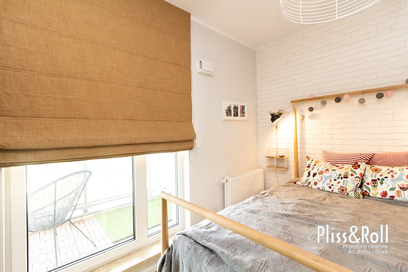 Beżowa, płócienna osłona okienna w stylu rzymskim, częściowo zwinięta, widoczna na tle jasnego pokoju z balkonem i białej ceglanej ściany.