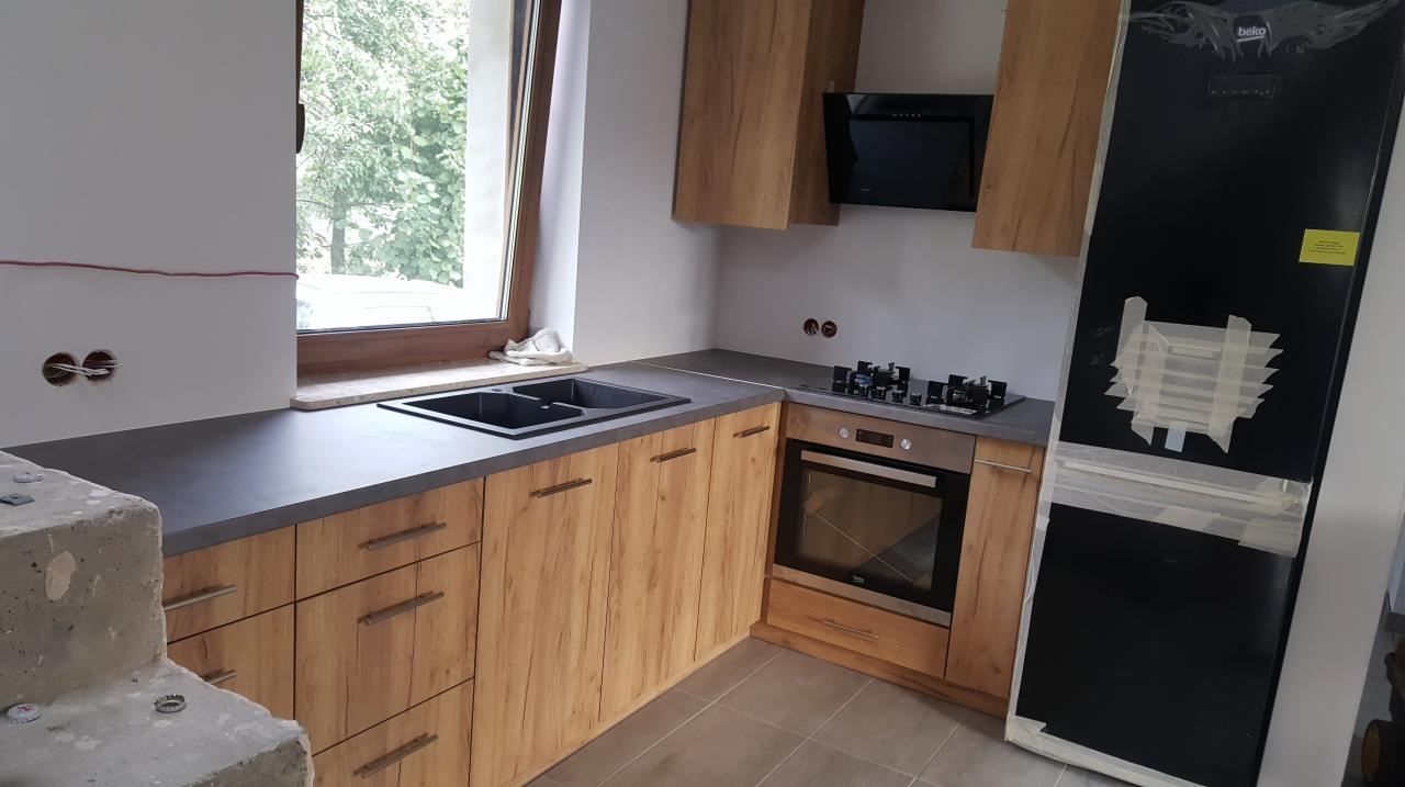 Montaż nowej kuchni z drewnianymi frontami i szarym blatem, widoczna płyta grzewcza, piekarnik oraz czarna lodówka zabezpieczona taśmą malarską.