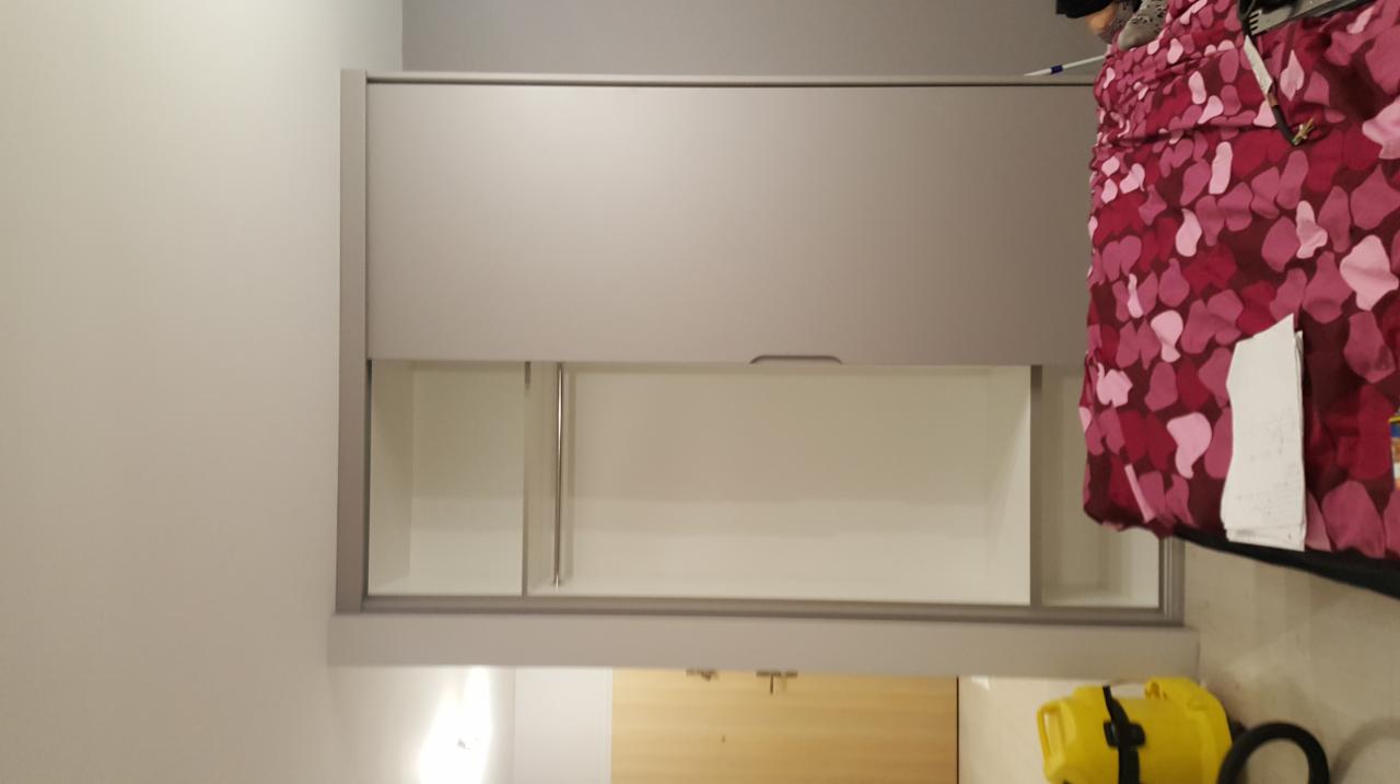 Szara szafa przesuwna z częściowo otwartymi drzwiami, widoczne półki wewnątrz, obok łóżko z bordową pościelą w kwiaty, na podłodze żółty pojemnik.