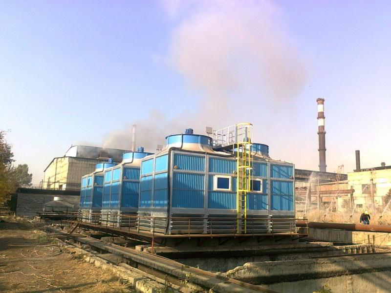 Rząd czterech niebieskich, przemysłowych wież chłodniczych z żółtymi schodami, umieszczonych na betonowej platformie, z kominem w tle i parą unoszącą się w powietrzu.