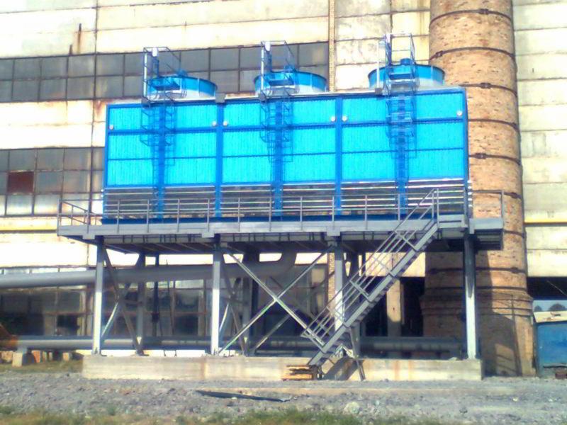 Instalacja chłodnicza z trzema wentylatorami na platformie stalowej, widoczna klatka schodowa i komin ceglany w tle, industrialny krajobraz.