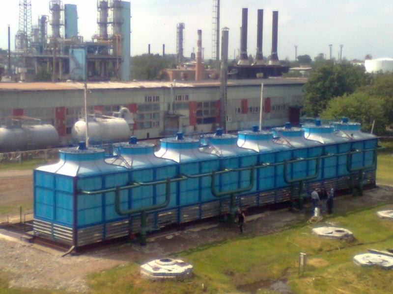 Instalacja chłodnicza z sześcioma modułami chłodniczymi na tle zakładu przemysłowego z kominami i zbiornikami, widoczne rury i osoby w pobliżu instalacji.