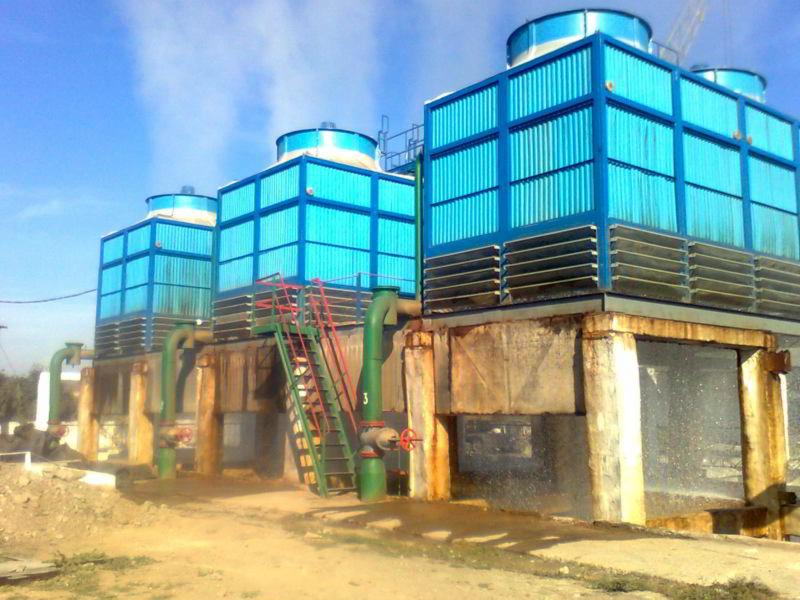 Kompleks czterech przemysłowych wież chłodniczych z niebieską obudową kratową, połączonych zielonymi rurami i schodami z czerwoną poręczą, z widoczną parą wydobywającą się z górnych otworów, na tle...