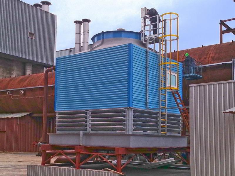 Montaż lub serwis przemysłowej wieży chłodniczej z niebieskimi panelami, dwóch pracowników na drabinie i podnośniku, na tle zabudowań przemysłowych z kominami.