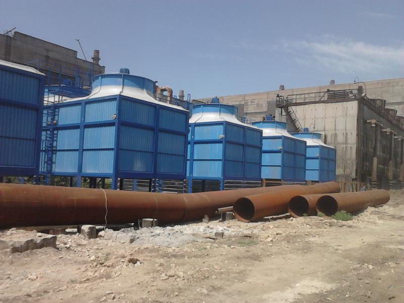 Instalacja chłodnicza: Cztery niebieskie wieże chłodnicze z rurami w rdzawym kolorze na placu budowy, z betonowym budynkiem przemysłowym w tle.