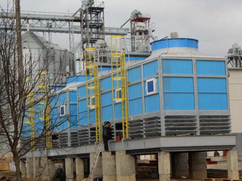 Instalacja chłodnicza przemysłowa z niebieskimi obudowami, żółtymi drabinkami i pracownikiem na drabinie, na tle silosów zbożowych