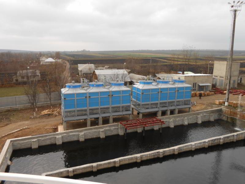 Dwa niebieskie agregaty chłodnicze wieżowe na betonowych fundamentach, widoczne z góry, z otwartym zbiornikiem wodnym i elementami konstrukcyjnymi w tle wiejskiego krajobrazu.