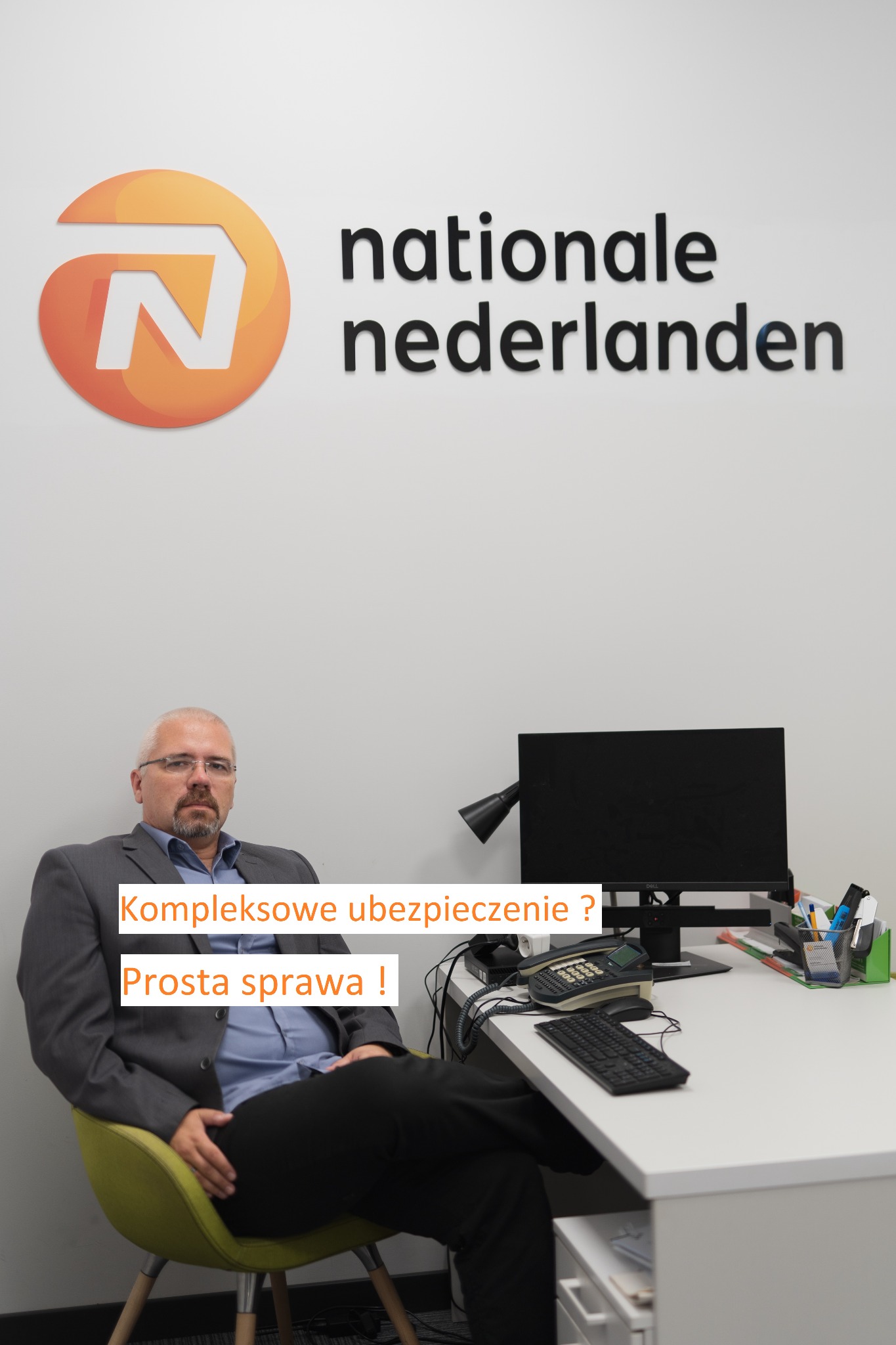 Przedstawiciel Nationale Nederlanden siedzący na krześle w biurze, za nim logo firmy, na biurku telefon stacjonarny, monitor i przybory biurowe.