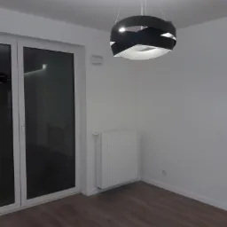 Pomalowane na biało wnętrze pokoju z drewnianą podłogą, grzejnikiem i nowoczesną lampą wiszącą w kształcie czarnej wstęgi, widok na białe drzwi balkonowe.