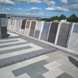 Ekspozycja różnorodnych płyt chodnikowych i elewacyjnych w punkcie sprzedaży w Kowale, z widocznymi wzorami ułożonymi na podłożu oraz pionowym panelem prezentującym próbki materiałów.