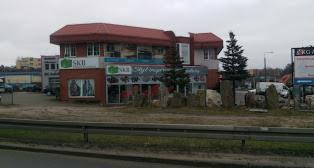 Dwukondygnacyjny budynek z czerwonym dachem, prawdopodobnie sklep lub biuro, z reklamami na fasadzie, otoczony kamieniami dekoracyjnymi i zielenią, widoczny zza barierek przy drodze.