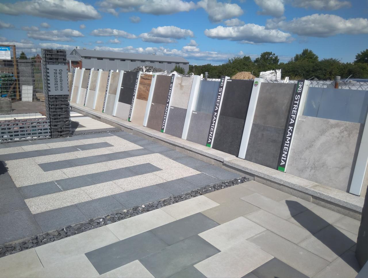 Ekspozycja różnorodnych płyt chodnikowych i elewacyjnych w punkcie sprzedaży w Kowale, z widocznymi wzorami ułożonymi na podłożu oraz pionowym panelem prezentującym próbki materiałów.