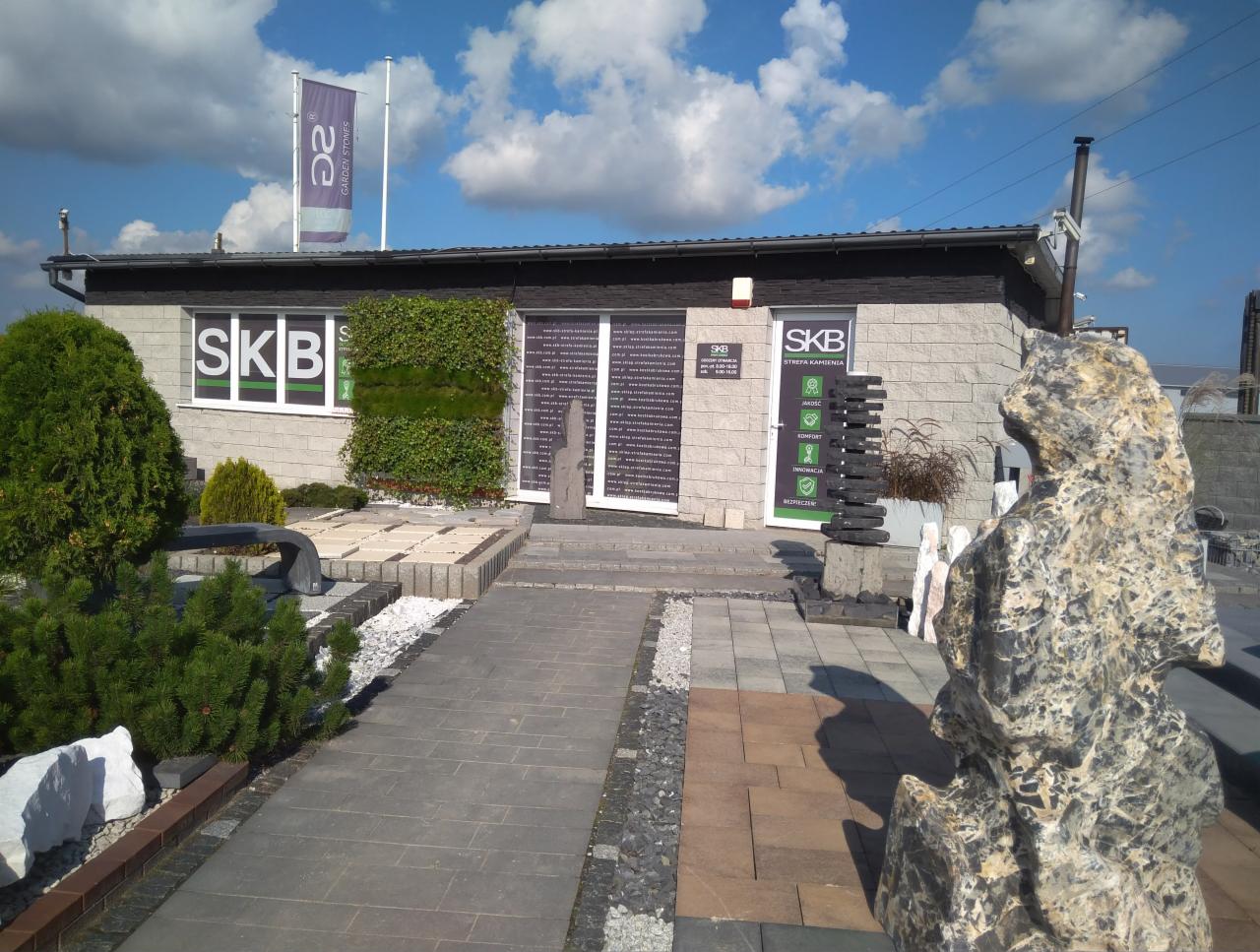 Ekspozycja składu budowlanego SKB z kostką brukową, elementami dekoracyjnymi z kamienia oraz zielenią przed budynkiem w słoneczny dzień.