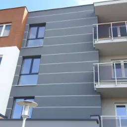 Nowoczesny budynek wielorodzinny z balkonami i elewacją w odcieniach bieli, brązu i szarości, z poziomymi pasami na szarej części fasady.