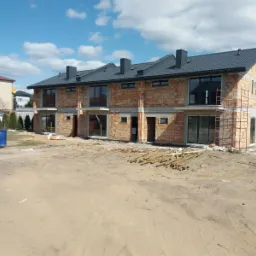 Surowy stan szeregowca z cegły ceramicznej, z widocznymi rusztowaniami i stertą drewna na placu budowy, pod błękitnym niebem z białymi chmurami.