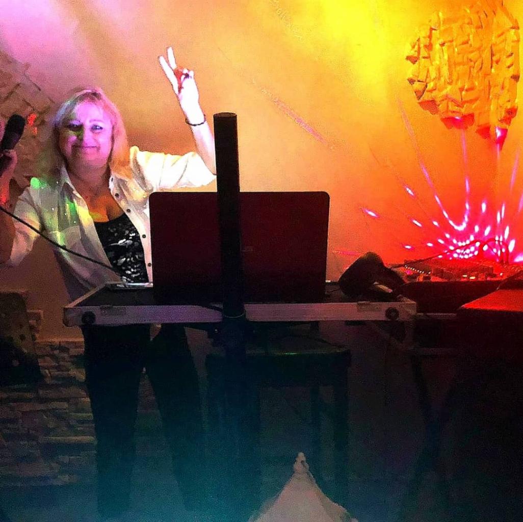 Uśmiechnięta blond DJ-ka z mikrofonem w ręku i znakiem zwycięstwa przed laptopem i sprzętem DJ-skim, oświetlona kolorowymi światłami na imprezie.