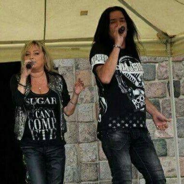 Dwoje wokalistów, kobieta z blond włosami i mężczyzna z długimi czarnymi włosami, śpiewają do mikrofonów na scenie. Kobieta ubrana w czarną koszulkę z napisem 'SUGAR CAN'T COMPETE' i błyszczącą...