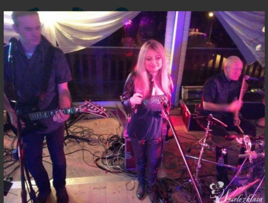 Zespół muzyczny grający na weselu: gitarzysta z gitarą elektryczną po lewej, wokalistka z mikrofonem pośrodku i perkusista z pałeczkami po prawej, scena udekorowana białymi materiałami.