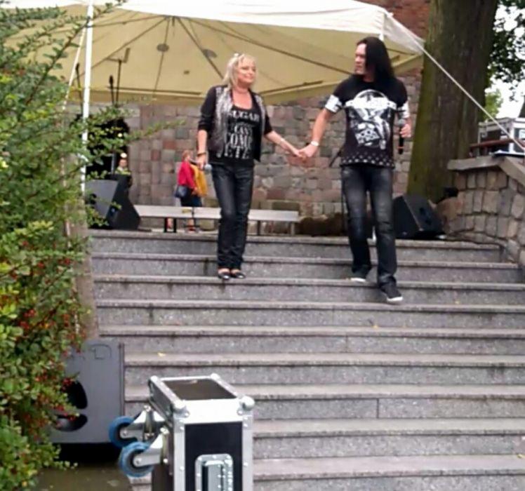 Dwoje wykonawców w czarnych strojach schodzi po schodach trzymając się za ręce, scena z nagłośnieniem i biały namiot w tle, widok z dołu.