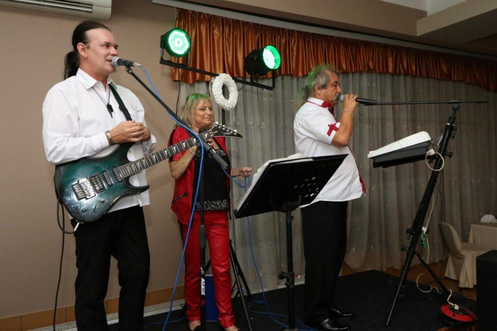 Zespół muzyczny w trakcie występu: gitarzysta z zielono-niebieską gitarą elektryczną śpiewa do mikrofonu, obok kobieta gra na keytarze, a po prawej stronie wokalista śpiewa do mikrofonu, na scenie...