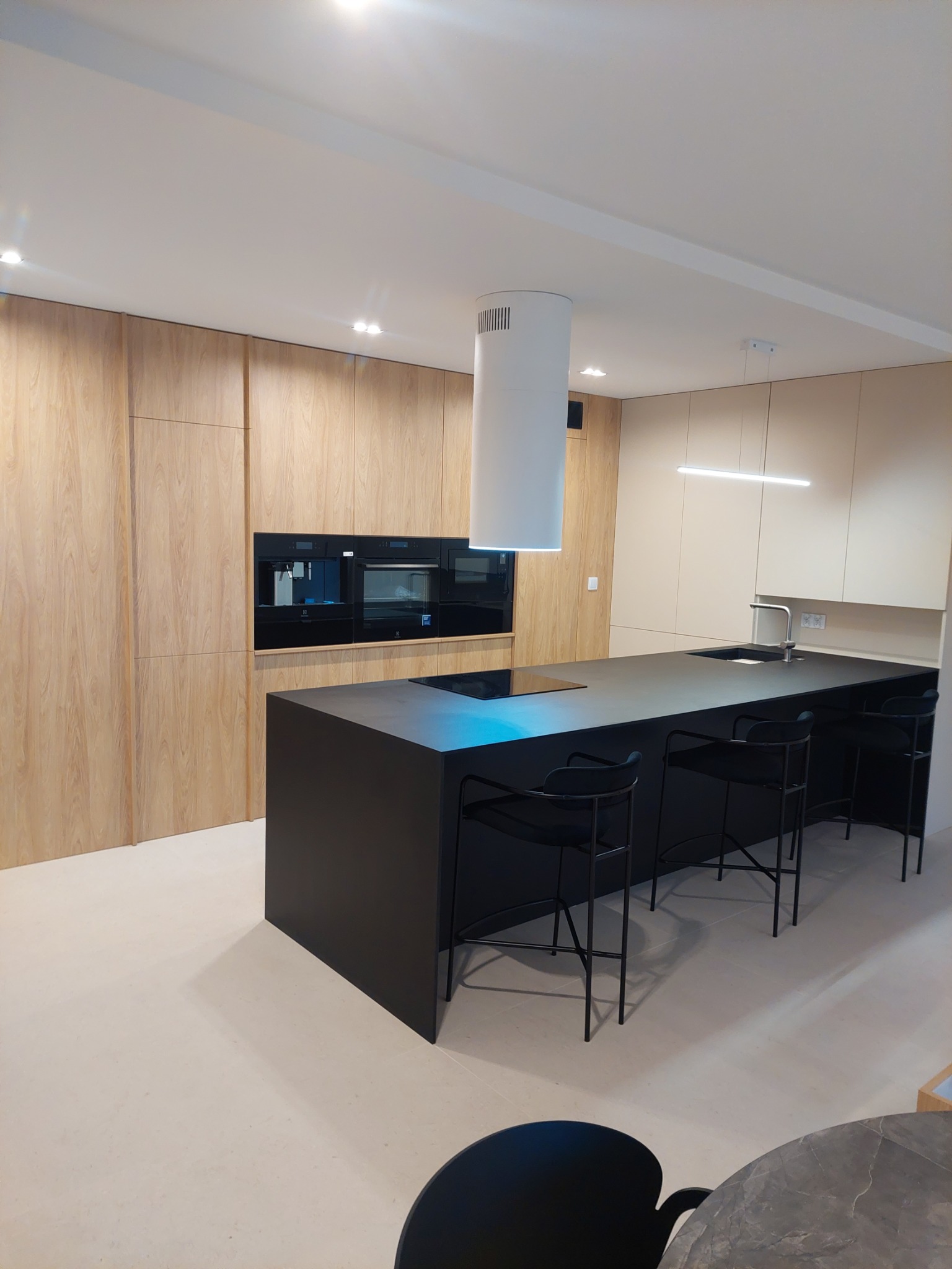 Nowoczesna kuchnia z drewnianymi szafkami, czarnym blatem wyspy kuchennej i białym okrągłym okapem; w tle zabudowane sprzęty AGD.