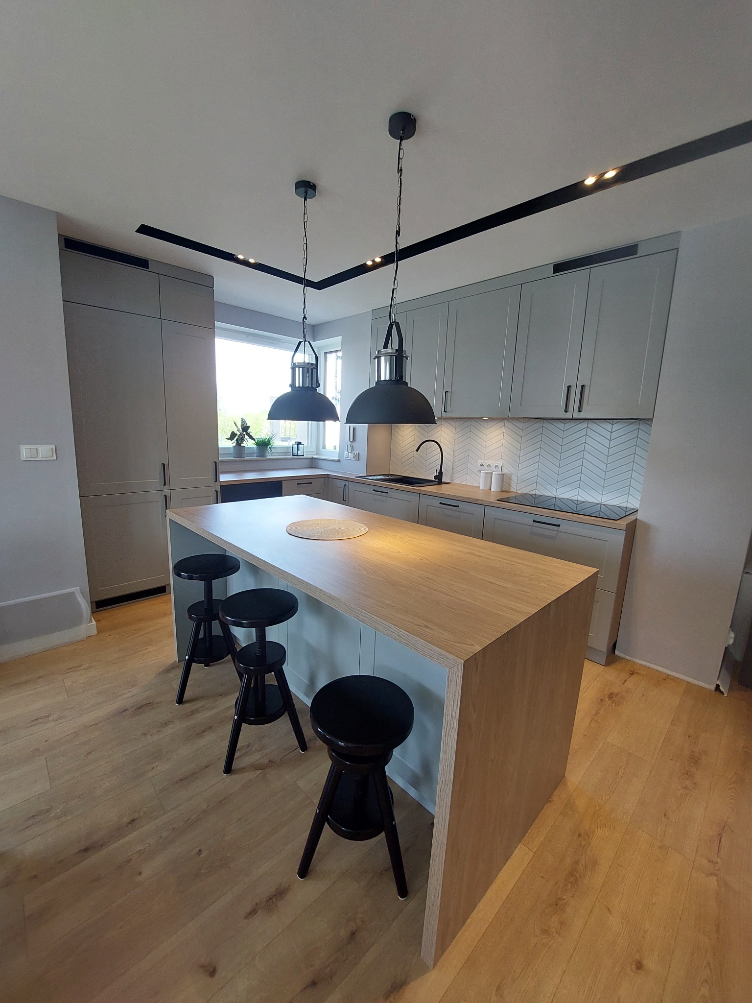 Szara kuchnia z drewnianym blatem wyspowym, trzema czarnymi stołkami barowymi i wiszącymi lampami w stylu industrialnym.
