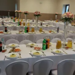 Sala bankietowa przygotowana na uroczystość, stoły nakryte białymi obrusami, zastawa, napoje, dania na talerzach, wysokie wazony z kwiatami, dekoracja w kształcie serca z kwiatów na ścianie...