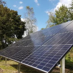 Czarny panel słoneczny ustawiony na metalowych wspornikach na tle zieleni i błękitnego nieba z chmurami.