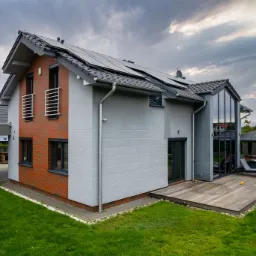 Nowoczesny dom z panelami słonecznymi na dachu, drewnianym tarasem i elementami cegły. Widok z zewnątrz na zadbany ogród i altanę.