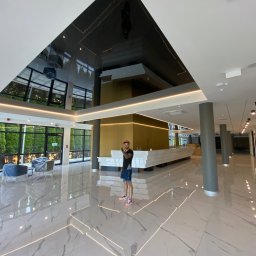 SHARP DESIGN Piotr Golecki - Błyszczący, czarny sufit napinany w przestronnym holu z marmurową podłogą, oświetleniem LED i osobą w tle, odbijający światło i otoczenie.