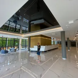 Błyszczący, czarny sufit napinany w przestronnym holu z marmurową podłogą, oświetleniem LED i osobą w tle, odbijający światło i otoczenie.