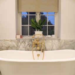 SHARP DESIGN Piotr Golecki - Elegancka, wolnostojąca wanna na tle marmurowej ściany. Złota armatura, roślina w doniczce na parapecie. Luksusowa łazienka po remoncie, detale wykończeniowe.